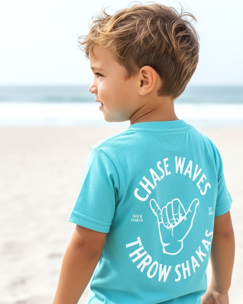 Child wearing a turquoise shirt with 'Chase Waves, Throw Shakas' text on a beach.